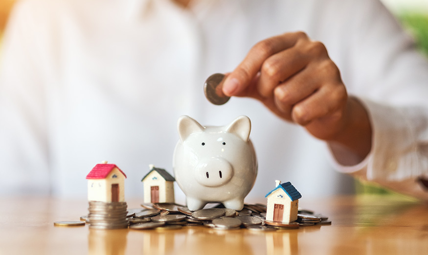Une femme place des pièces dans une tirelire, avec des maquettes de maisons posées sur une pile de pièces, illustrant le concept d'épargne pour l'achat immobilier.