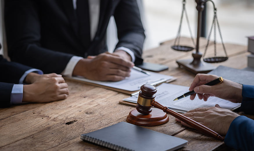Deux personnes assises face à un procureur s’apprêtant à signer un document, avec une balance de la justice et le marteau du juge posés sur son bureau.