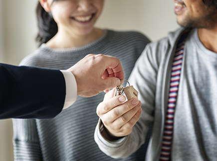 Un homme en costume tend une clé avec un porte clé en forme de maison à un couple qui se regarde en souriant.