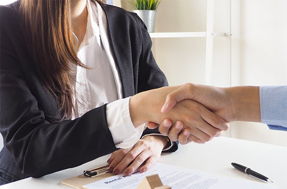 Une femme en costume assise à un bureau sert la main d’un homme en lui tendant un contrat
