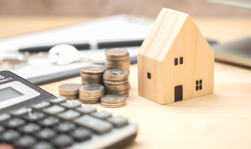 Une petite maison en bois à coté d'une calculatrice et de pièces de monnaie.